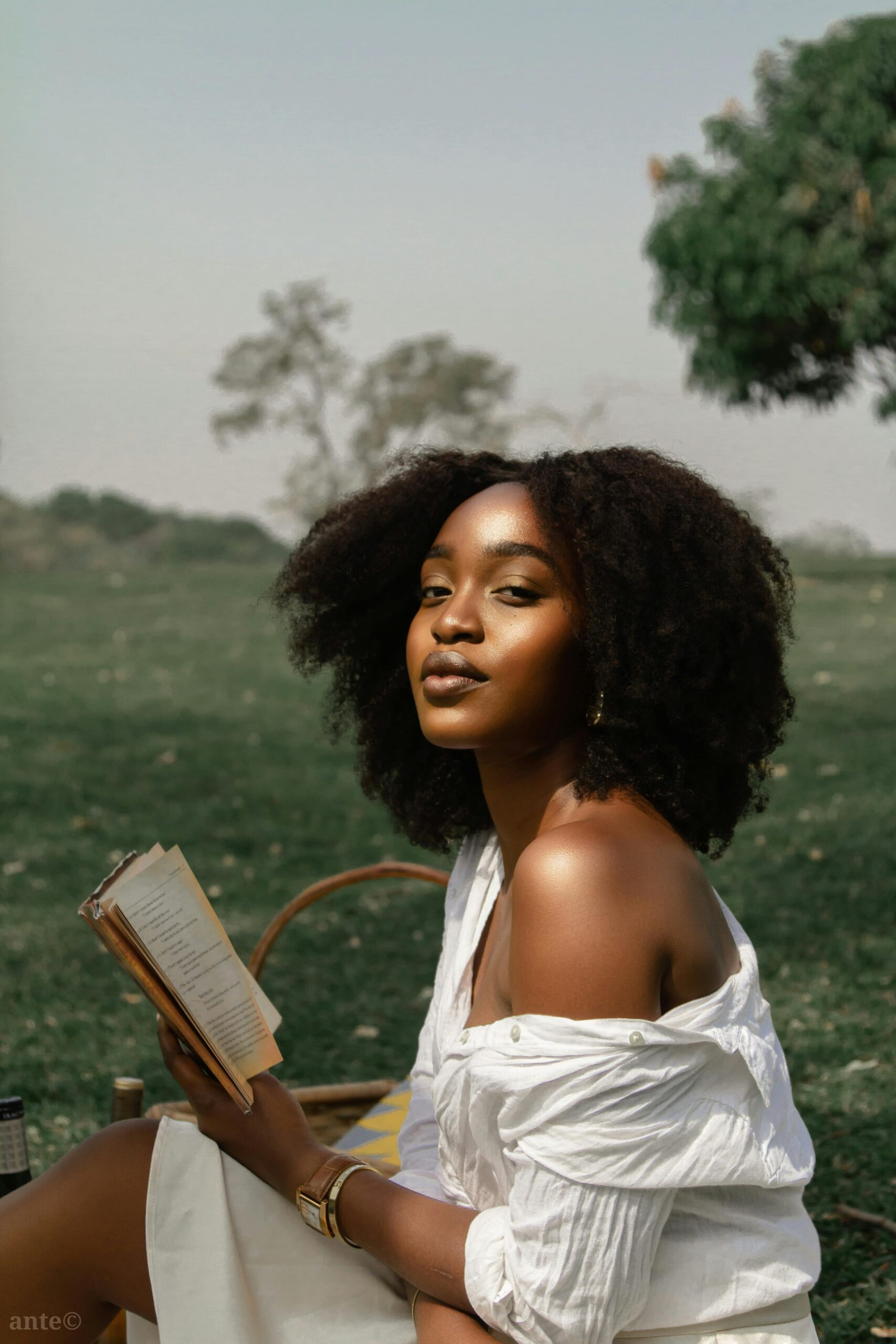 Picture of a Black girl reading her book in a field, looking at camera